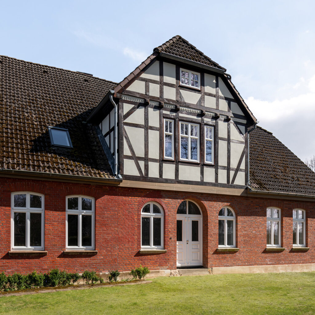 Straßenansicht des sanierten Landhauses in Holthusen mit Ziegelfassade, wiederhergestelltem Fachwerk-Zwerchhaus und neuer Eingangstür mit Rundbogen.