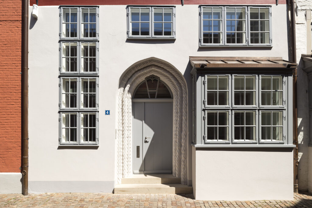 Sanierte historische Fassade eines Stadthauses mit heller Putzoberfläche, aufwendig gestaltetem gotischem Sandsteinportal, grauen Sprossenfenstern und modernem Erker in der Altstadt von Lüneburg.