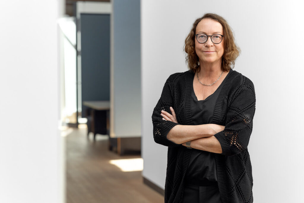 Dunkelhaarige Frau mit Brille und verschränkten Armen steht im Büroflur. Sie trägt ein schwarzes T-Shirt und eine schwarze Strickjacke.