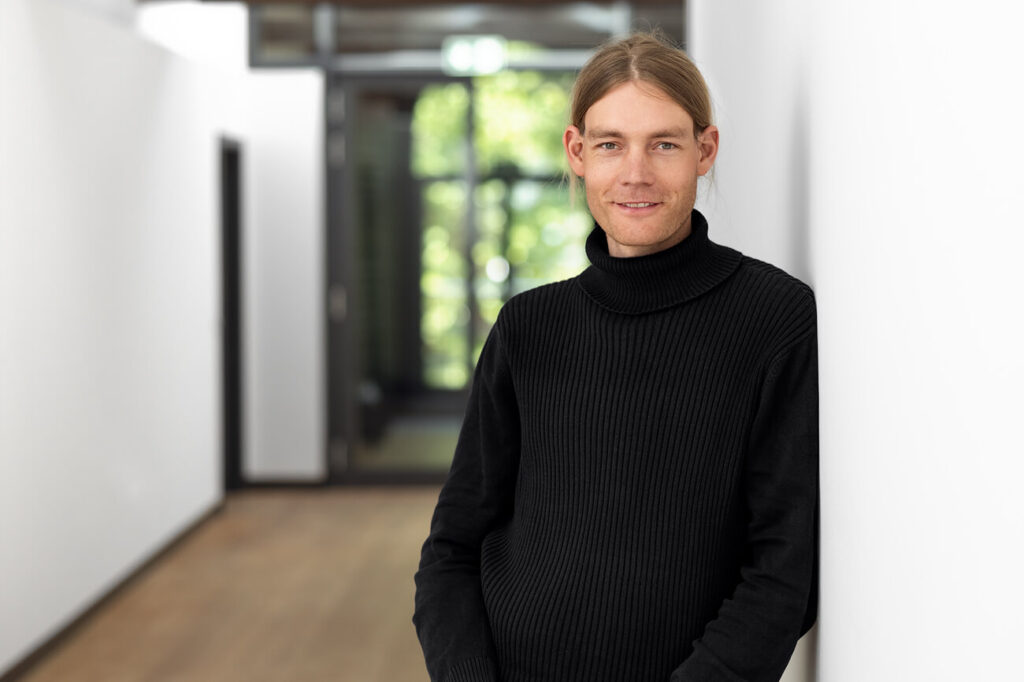Dunkelhaariger Mann steht an Wand angelehnt im Büroflur. Er trägt ein schwarzen Rollkragenpullover und lächelt.
