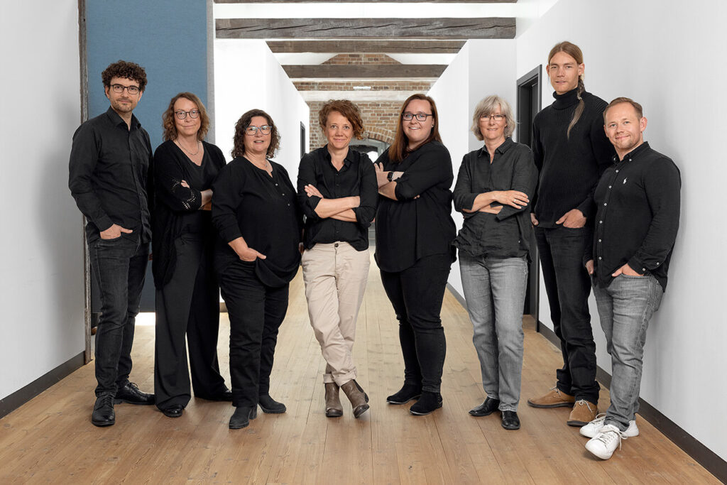 8-Köpfiges Team der Firma Saborosch, bestehend aus fünf Frauen und drei Männern, stehen zusammen als Gruppe in Reihe im Büroflur.
