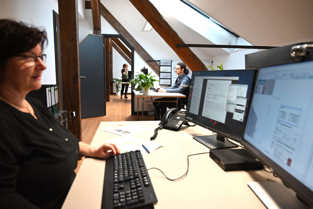 Zwei Frauen und ein Mann mittleren Alters im Büro, sie sind vertieft in ihre Arbeit am Schreibtisch und schauen auf den Monitor. Einer der beiden Frauen telefoniert stehend am Schreibtisch.